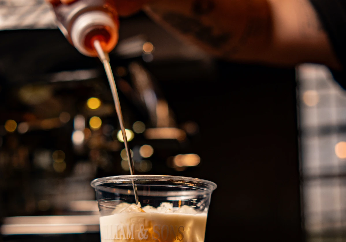 Um barista adicionando um xarope em um copo de bebida com gelo, destacando a técnica de preparo de bebidas.